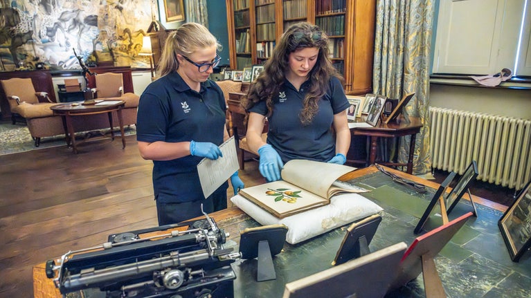 Two National Trust staff members, wearing navy blue polo shirts with the NT oak leaf logo, carrying out conservation work on a large, old book.